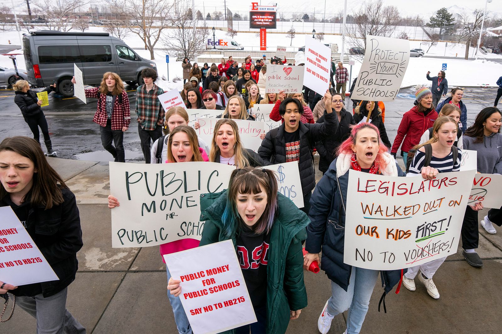 A group of students and adults stand outside holding signs opposing school vouchers. There is snow on the ground.