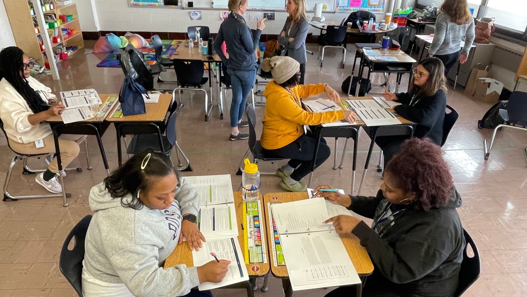 A photo of a school classroom shows adults working at individual desks with paper, pens and pencils. Several other adults are standing and talking or walking.